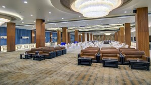 A spacious hall with rows of brown leather sofas and tables, a large chandelier, and a blue curtain backdrop. - The Bindal Hotel (Jamshedpur)