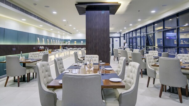 A modern restaurant with multiple dining tables and chairs, a central pillar, and a view of the city through large windows. - The Bindal Hotel (Jamshedpur)