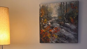 Een kamer met een schilderij van een boslandschap, een lamp en een mosterdgele stoel.