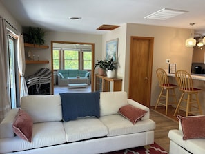 A living room with a sofa, a dining area with chairs, a kitchen, and a large window with a view.