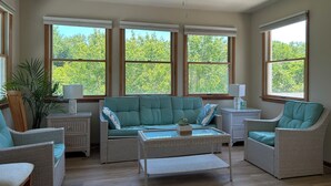 A room with a ceiling fan, large windows, wicker furniture, and a view of trees. - Spacious 3BR/3BA Beach House on Quiet Wooded Lot – Minutes from the Ocean (Kitty Hawk)