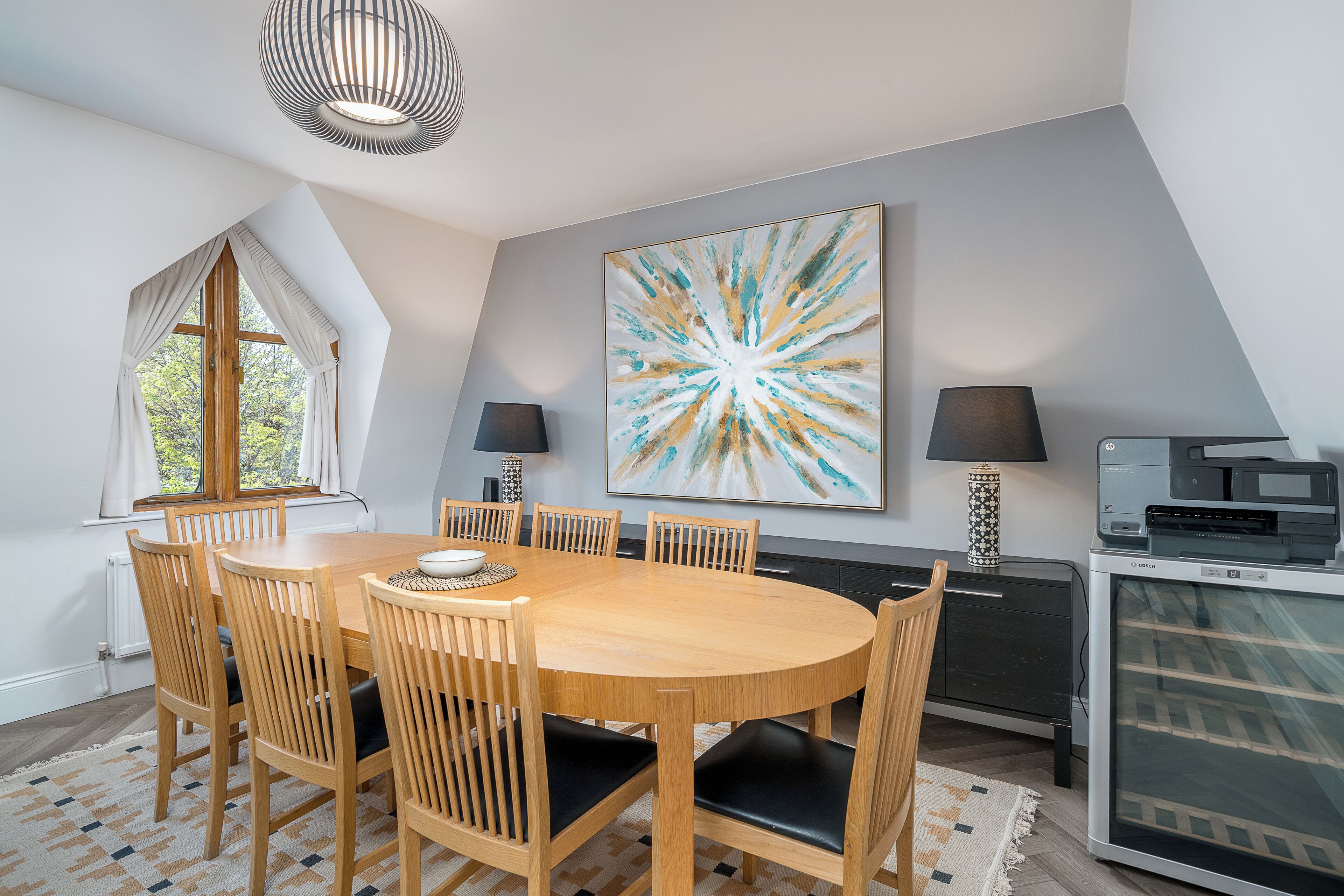 A dining area with a round wooden table, six chairs, two lamps, and a large abstract painting on the wall.