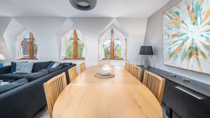 A modern dining area with a wooden table, chairs, and a black cabinet. There are large windows with white curtains and a view of greenery outside. - Chic 2BR Battersea Flat - Terrace & Pet-Friendly (London)