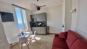 A modern living room with a red sofa, a dining area with white chairs, a kitchen with grey cabinets, and a large window with a view of the sea. - Vue mer - Monaco - clim (Cap-d'Ail)