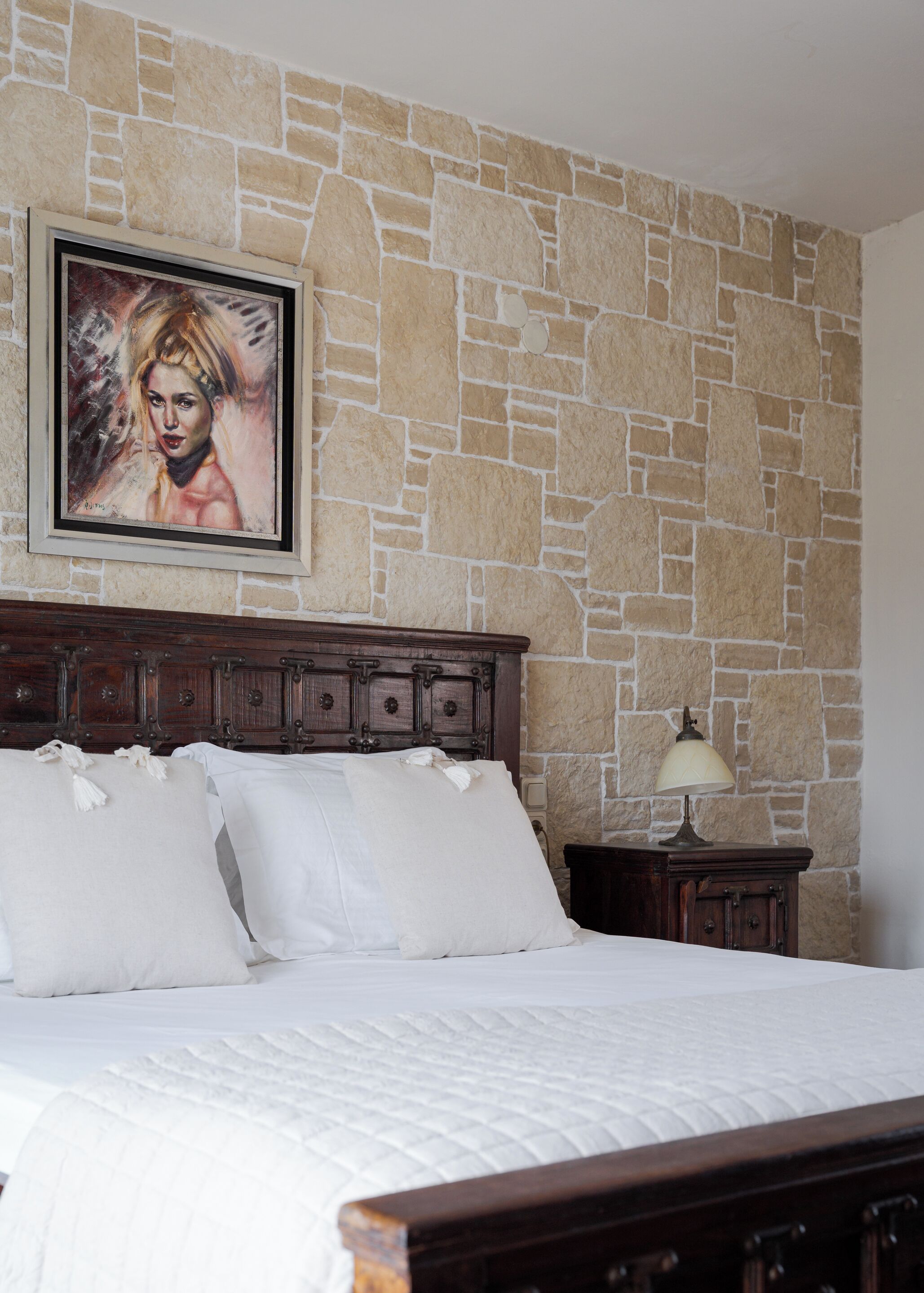Un dormitorio con pared de piedra, cabecera de madera, ropa de cama blanca y una obra enmarcada sobre la cama.