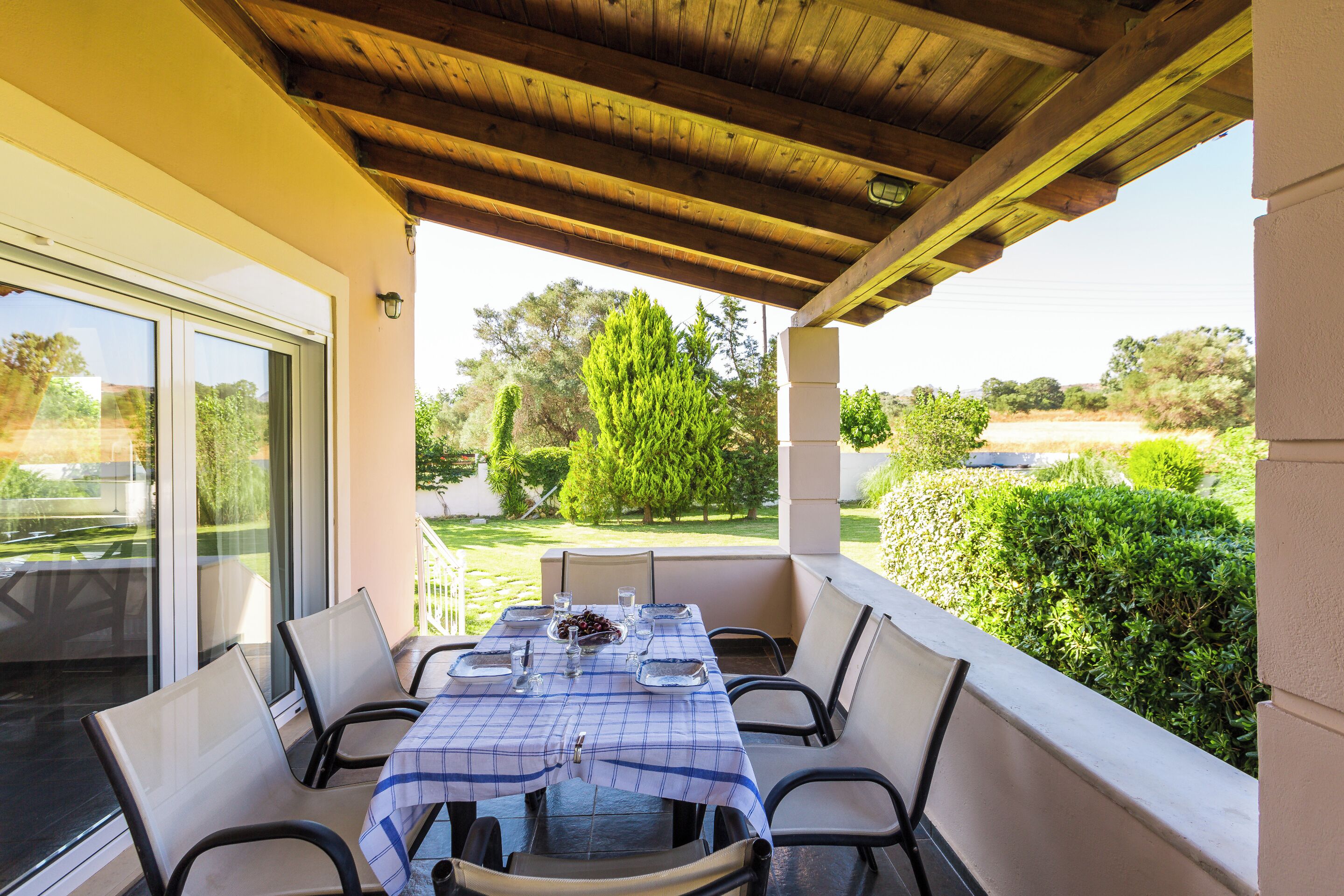 A covered outdoor dining area with a table set for four, surrounded by chairs, and a view of the garden and trees.