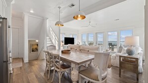 A spacious living area with a wooden dining table, white upholstered chairs, and a large window offering a view of the ocean. - Gulf Views with Hot Tub, Garage and Bikes (Santa Rosa Beach)