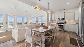 A spacious kitchen with white cabinetry, a dining area with a wooden table and white chairs, and a view of the ocean through large windows. - Gulf Views with Hot Tub, Garage and Bikes (Santa Rosa Beach)