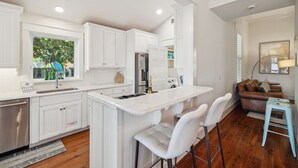 Una cocina moderna con armarios blancos, una isla central con taburetes y un refrigerador de acero inoxidable, y un ventanal con vistas a la vegetación.