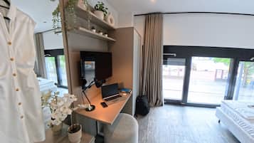 A modern hotel room with a desk, laptop, and a television. There are potted plants and a bed with a white comforter.