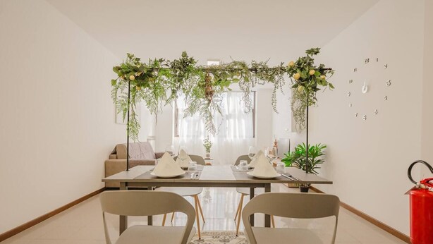 Zona pranzo con tavolo imbandito per quattro persone, circondata da sedie, un divano e un arco decorativo con piante.