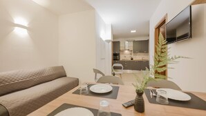 Un espace de vie compact comprenant un canapé, une table à manger pour deux personnes et une cuisine située à l’arrière-plan.