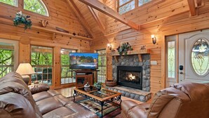 A log cabin interior with a fireplace, leather sofas, a television, and a stone fireplace surround. - Elk Rapids On Cosby Creek 3 Bedroom Cabin (Cosby)