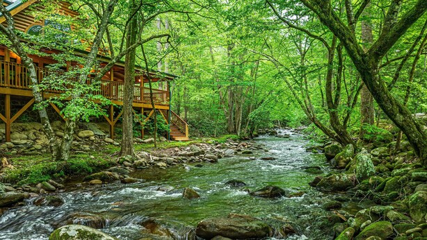 A wooden cabin with a deck overlooking a flowing stream in a forest. - Elk Rapids On Cosby Creek 3 Bedroom Cabin (Cosby)