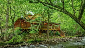 A wooden cabin with a deck and stairs, surrounded by a forest and a flowing stream. - Elk Rapids On Cosby Creek 3 Bedroom Cabin (Cosby)