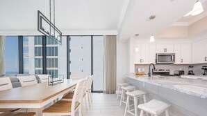 A modern kitchen with white cabinetry, a large island with bar stools, and a dining area with a long table and chairs. - Phoenix Gulf Shores Ii 1501 4 Bedroom Condo (Gulf Shores)
