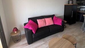 Un salon moderne avec un canapé noir, des coussins roses, une table basse ronde en bois et une petite table d’appoint avec une lampe.