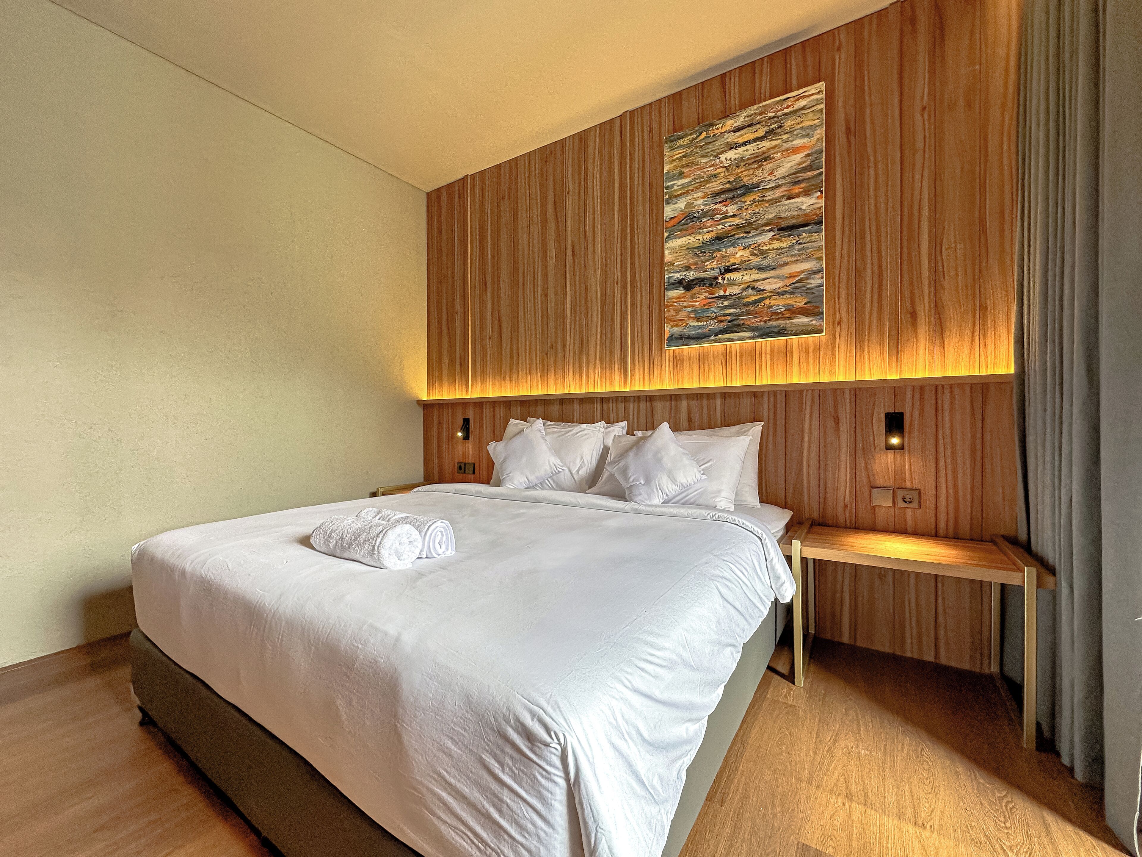 Una cama bien hecha con sábanas blancas y toallas enrolladas, un cabecero de madera con un cuadro y una mesita de noche con una lámpara.
