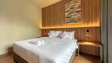 Una cama bien hecha con sábanas blancas y toallas enrolladas, un cabecero de madera con un cuadro y una mesita de noche con una lámpara.