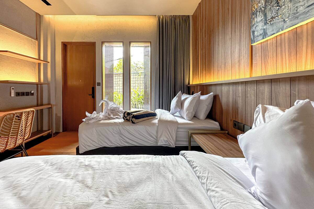 Habitación de hotel con dos camas, una zona de estar con una silla, un cabecero de madera y un amplio ventanal con vistas a zonas verdes.