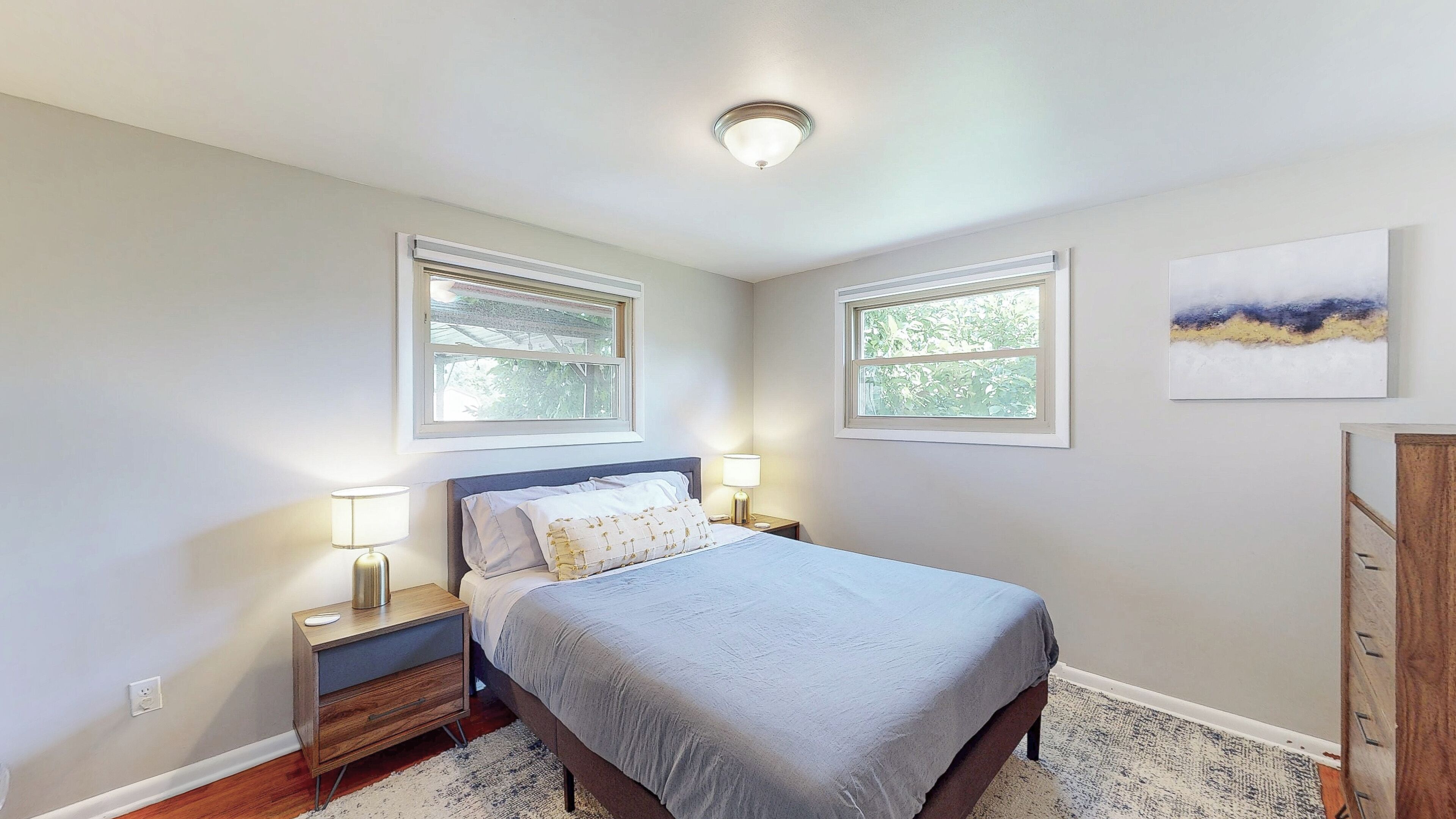 A bedroom with a bed, two bedside tables with lamps, a ceiling light, two windows, and a painting on the wall.