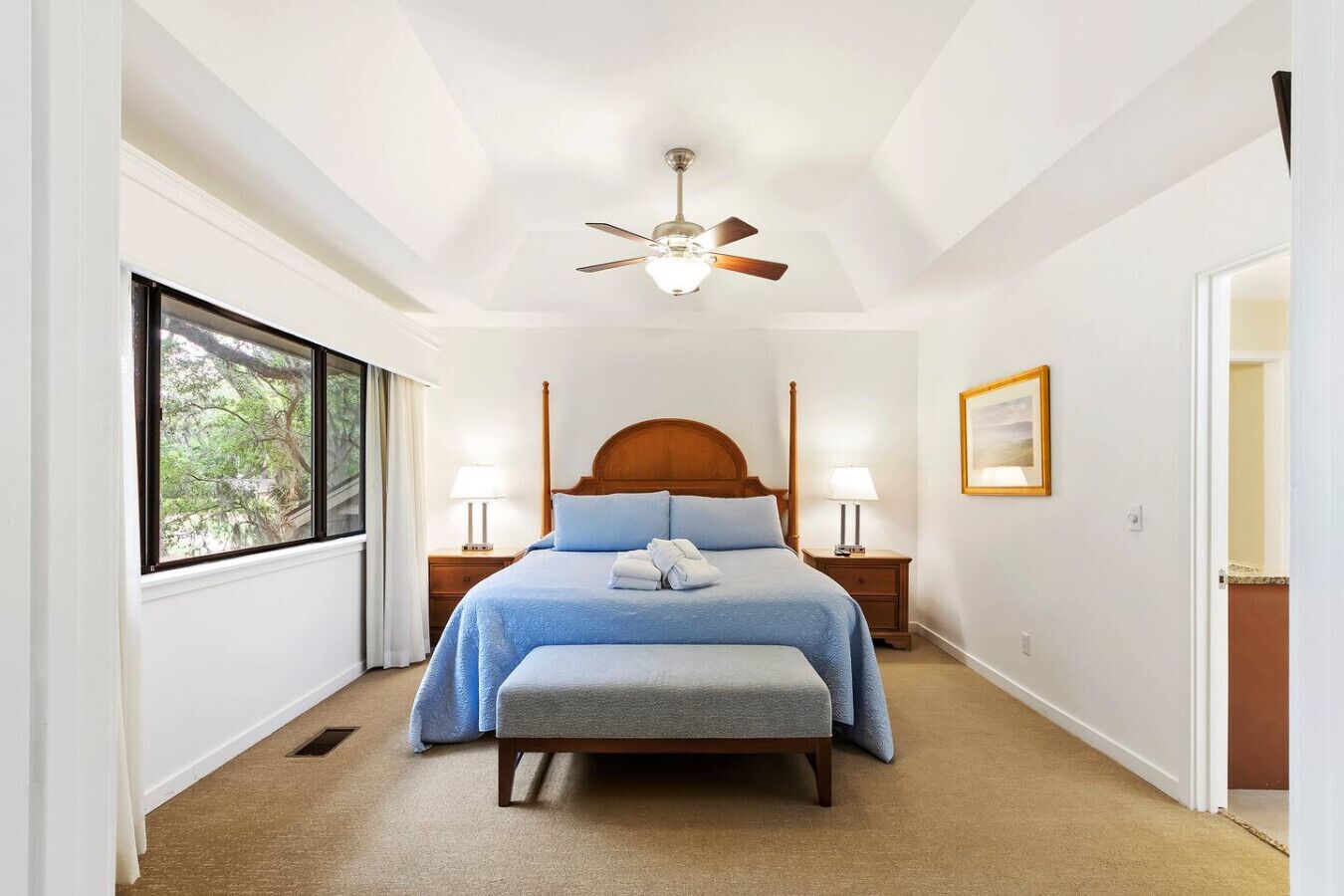 A bedroom with a single bed, a ceiling fan, bedside lamps, a window with a view of trees, and a framed picture on the wall.
