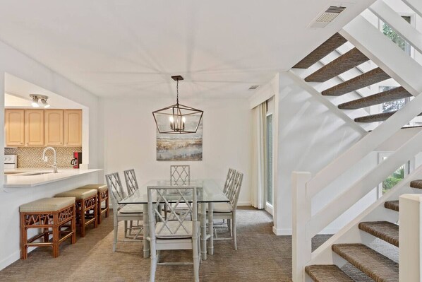 A spacious dining area with a glass table and chairs, a modern chandelier, and a staircase with wooden steps.