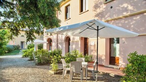 An outdoor seating area with tables and chairs under umbrellas, surrounded by greenery and a building with arched windows. - Resort with pool in Riparbella  (Riparbella)