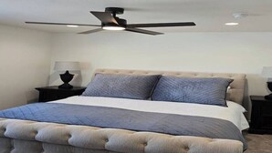 Een slaapkamer met een gestoffeerd hoofdbord, een bed met een gewatteerde spreidek en een plafondventilator, plus twee bedlampen.
