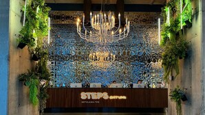 Zona de recepción con candelabro, mostrador de recepción y pared decorada con plantas y baldosas reflectantes.