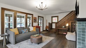 Ein Wohnzimmer mit grauem Sofa, Ottoman und einer Chandelier. Eine Treppe mit Holzwerteln und einem an der Wand befestigten Fernseher.