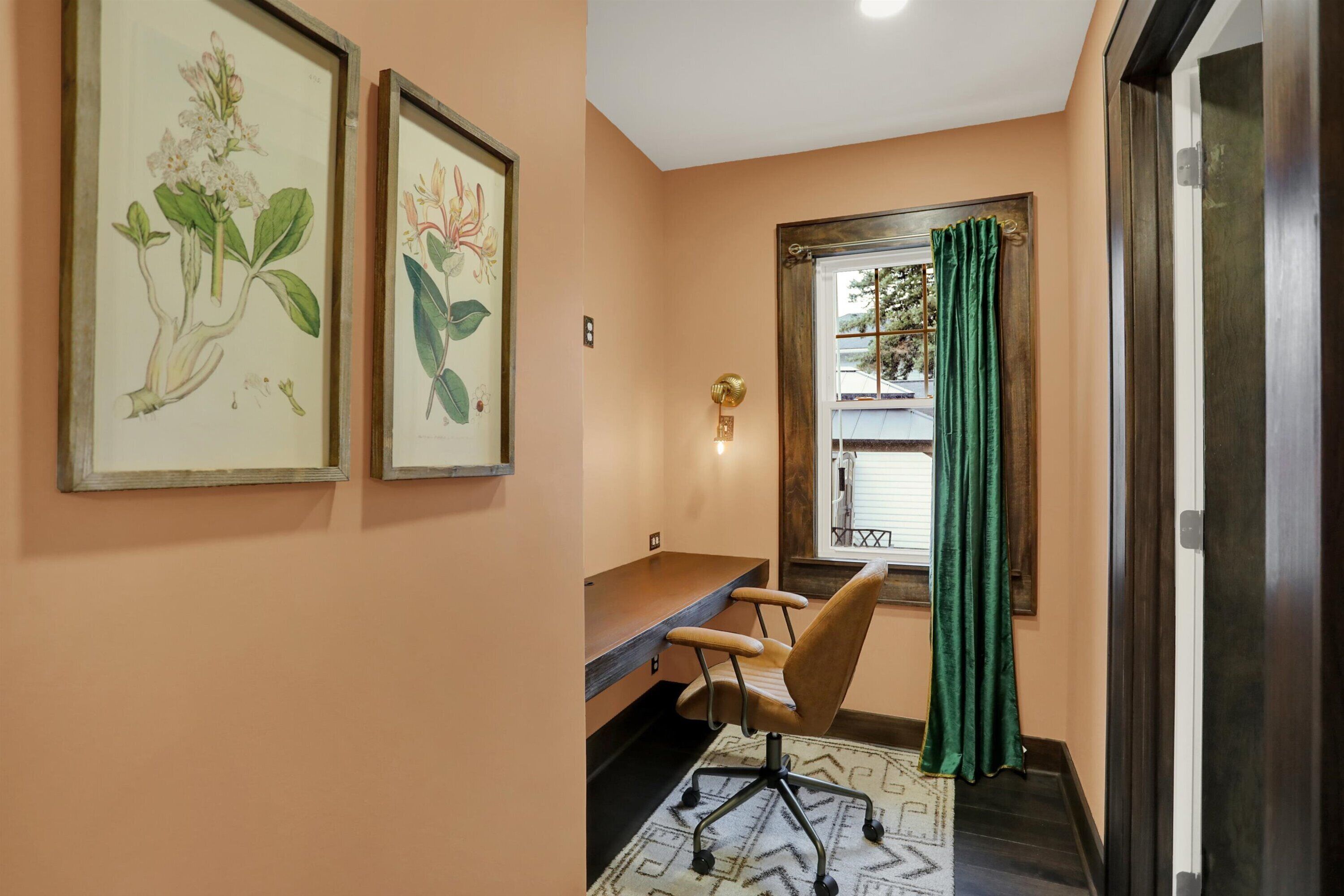 A room with a desk, chair, framed botanical illustrations, a window with a curtain, and a door.