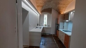 Una cuina estreta i volta amb una illa central, mobles blanqus i terra de rajola de pedra.
