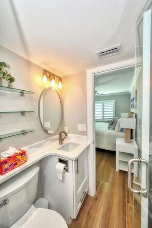 A compact bathroom with a toilet, sink, and mirror. There are glass shelves above the sink and a small bedside table with a lamp in the adjacent bedroom. - Seaside Resort Unit 309 (Fort Myers Beach)