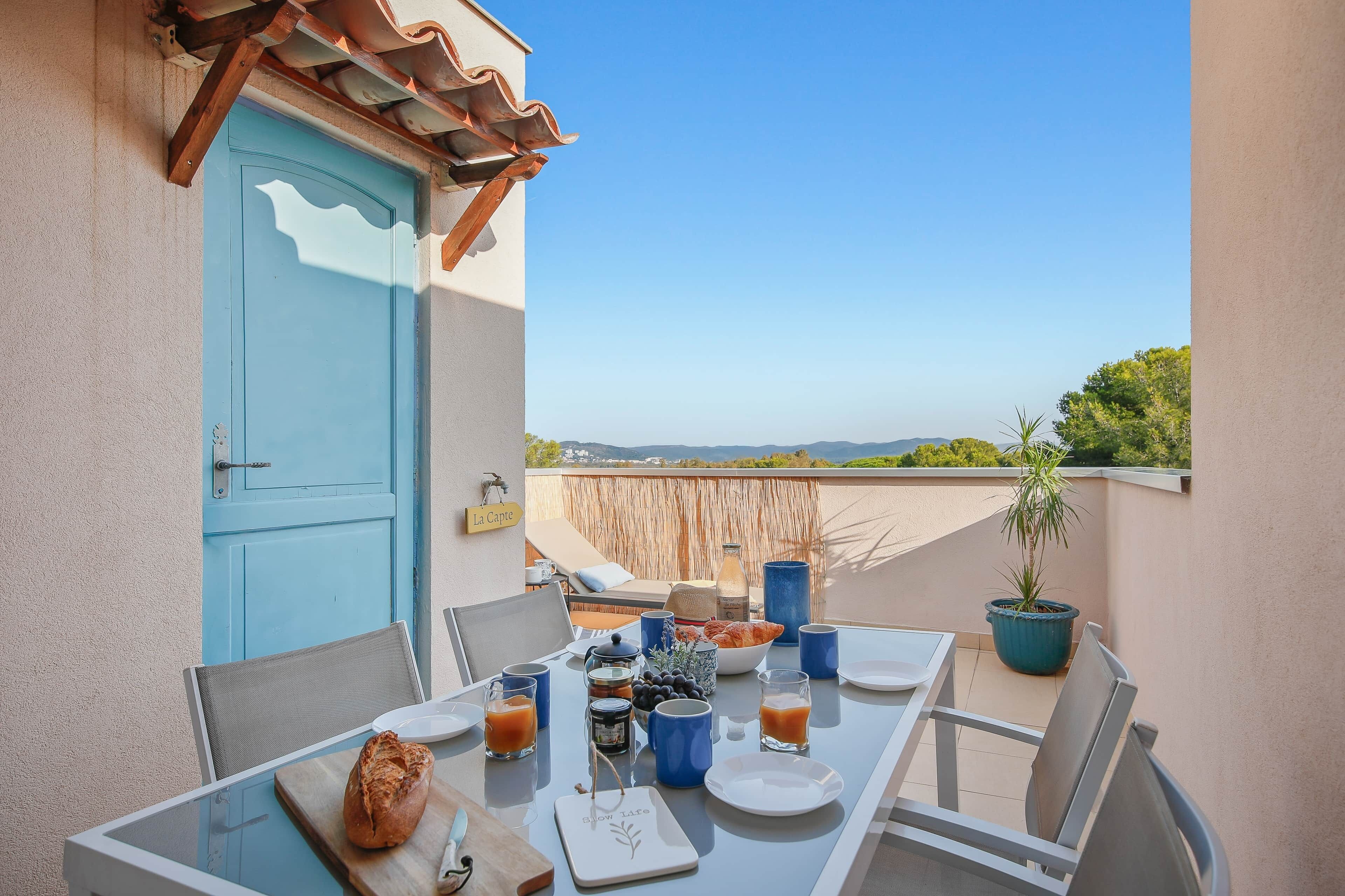 A balcony with a table set for breakfast, a blue door, and a view of the landscape.