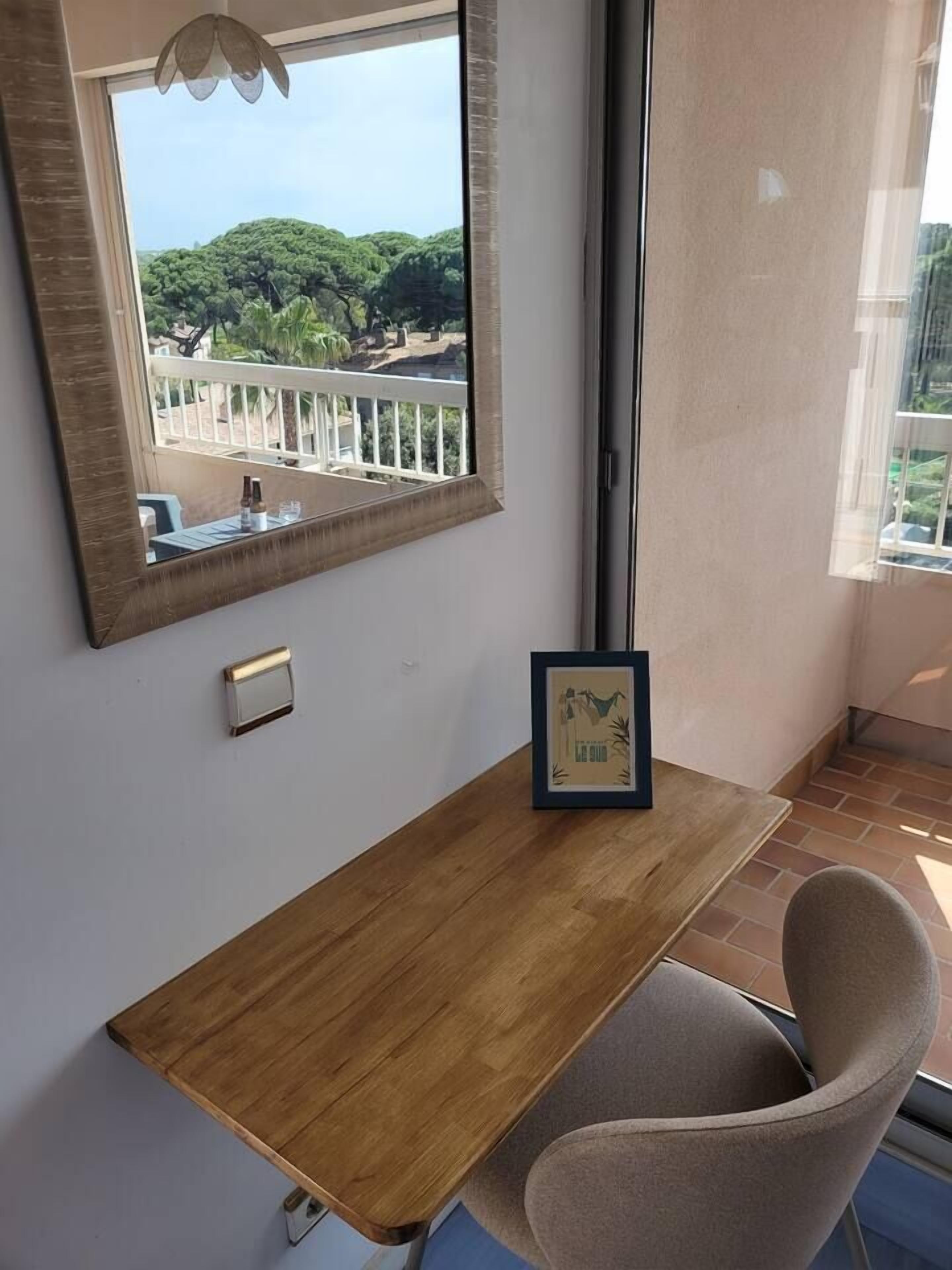 Een kamer met een houten bureau, een stoel, een ingelijst kunstwerk en een raam met uitzicht op bomen en een balkon.