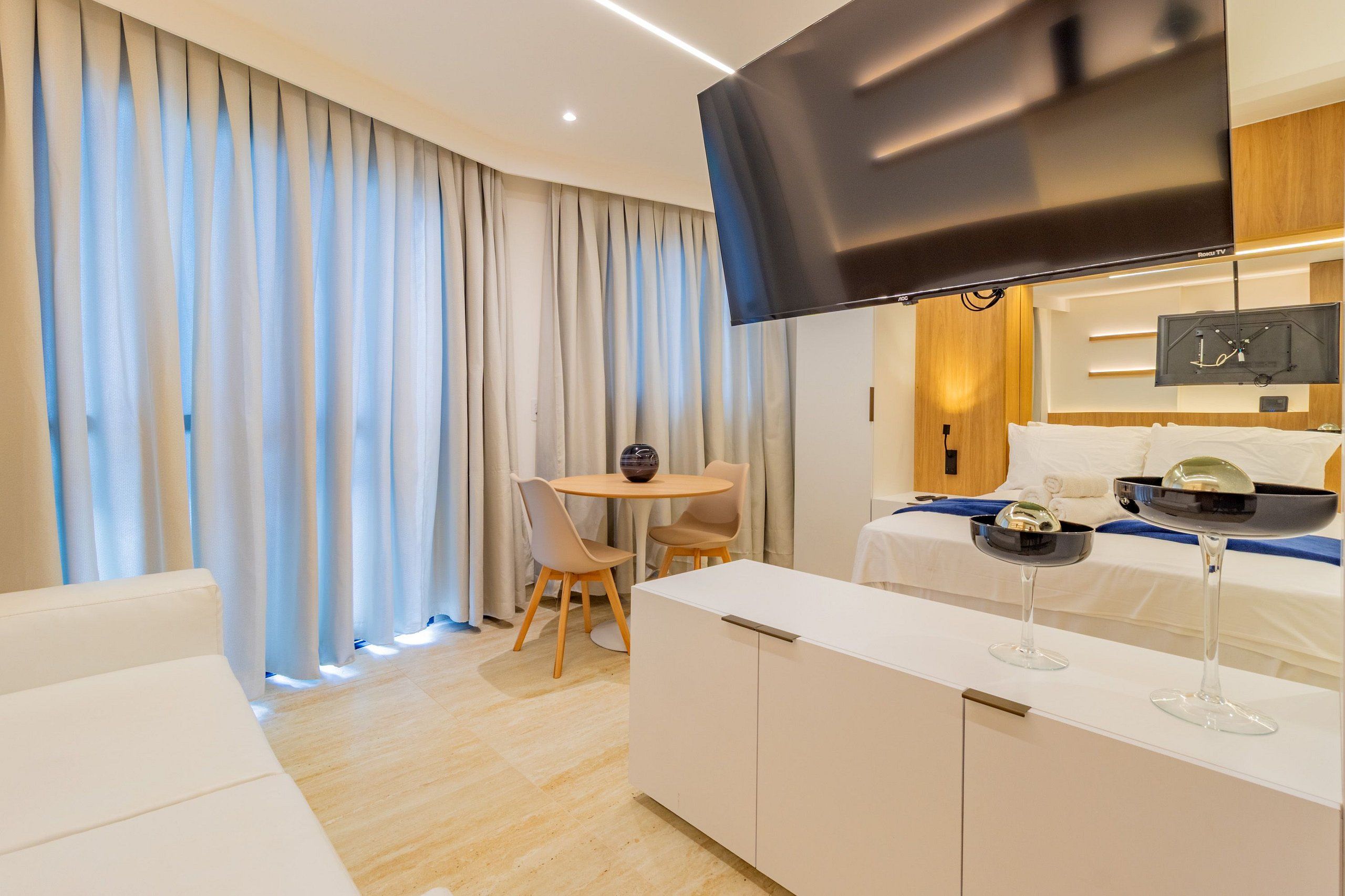 A modern hotel room with a flat-screen TV, a dining area with a table and chairs, and a bed with a TV on the headboard.