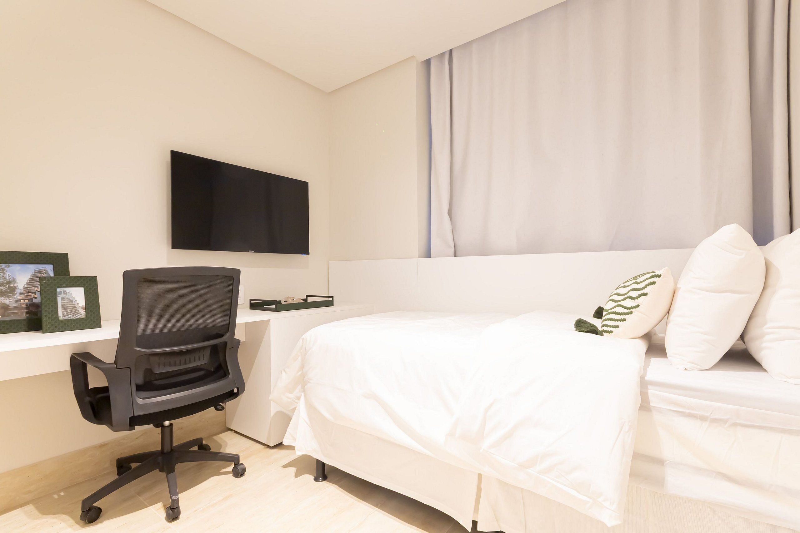 A bedroom with a bed, a desk with a chair, a flat-screen TV mounted on the wall, and a window with curtains.