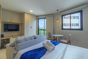 Ein Hotelzimmer mit Bett, Fernseher, Schreibtisch, Stuhl und einem Balkon mit Blick auf Gebäude.