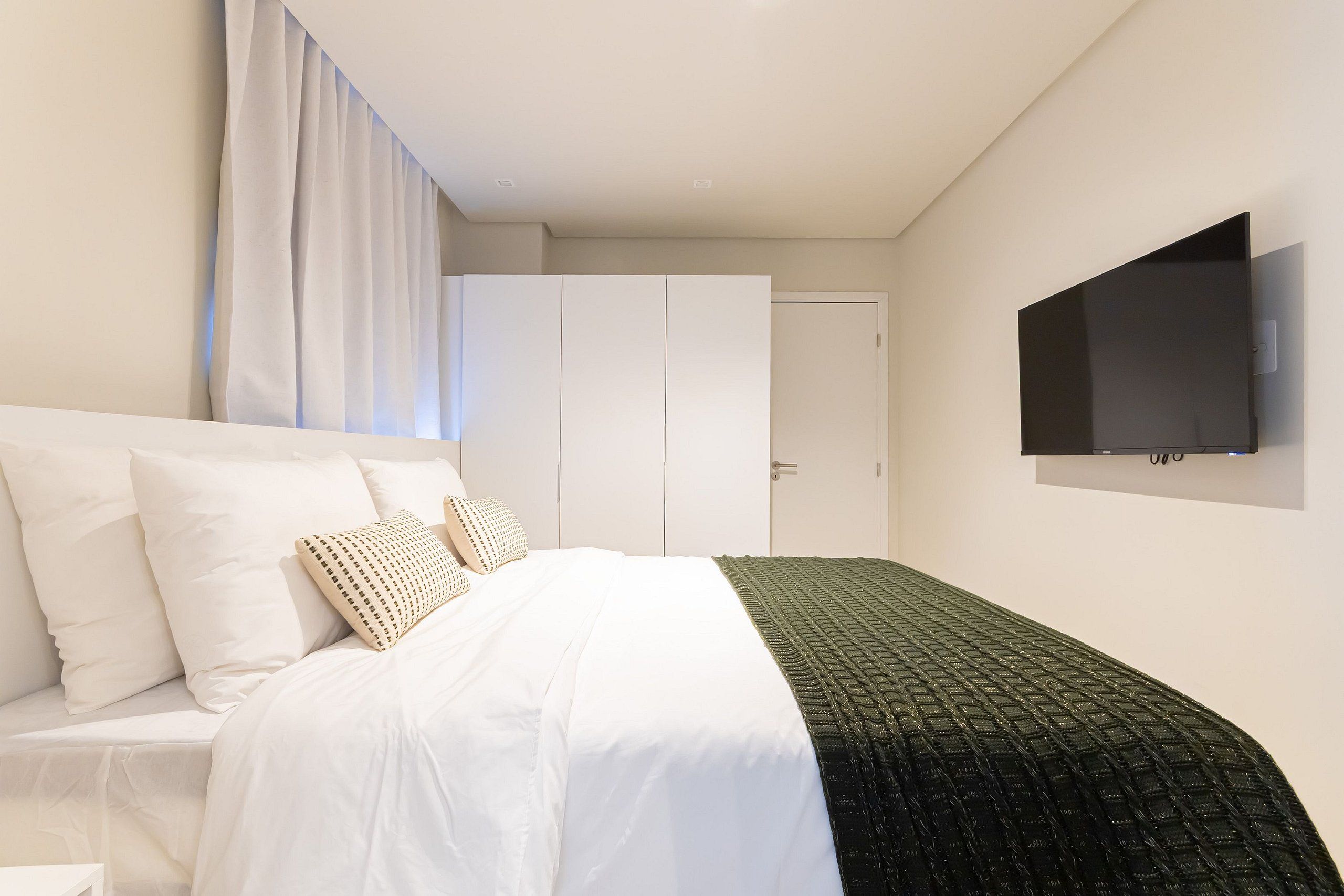 A bedroom with a bed, white curtains, a TV on the wall, and a wardrobe.