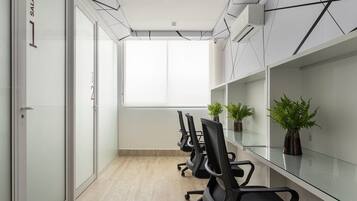 A conference room with glass walls, a white ceiling, and a large window. There are black office chairs and potted plants on the glass table.
