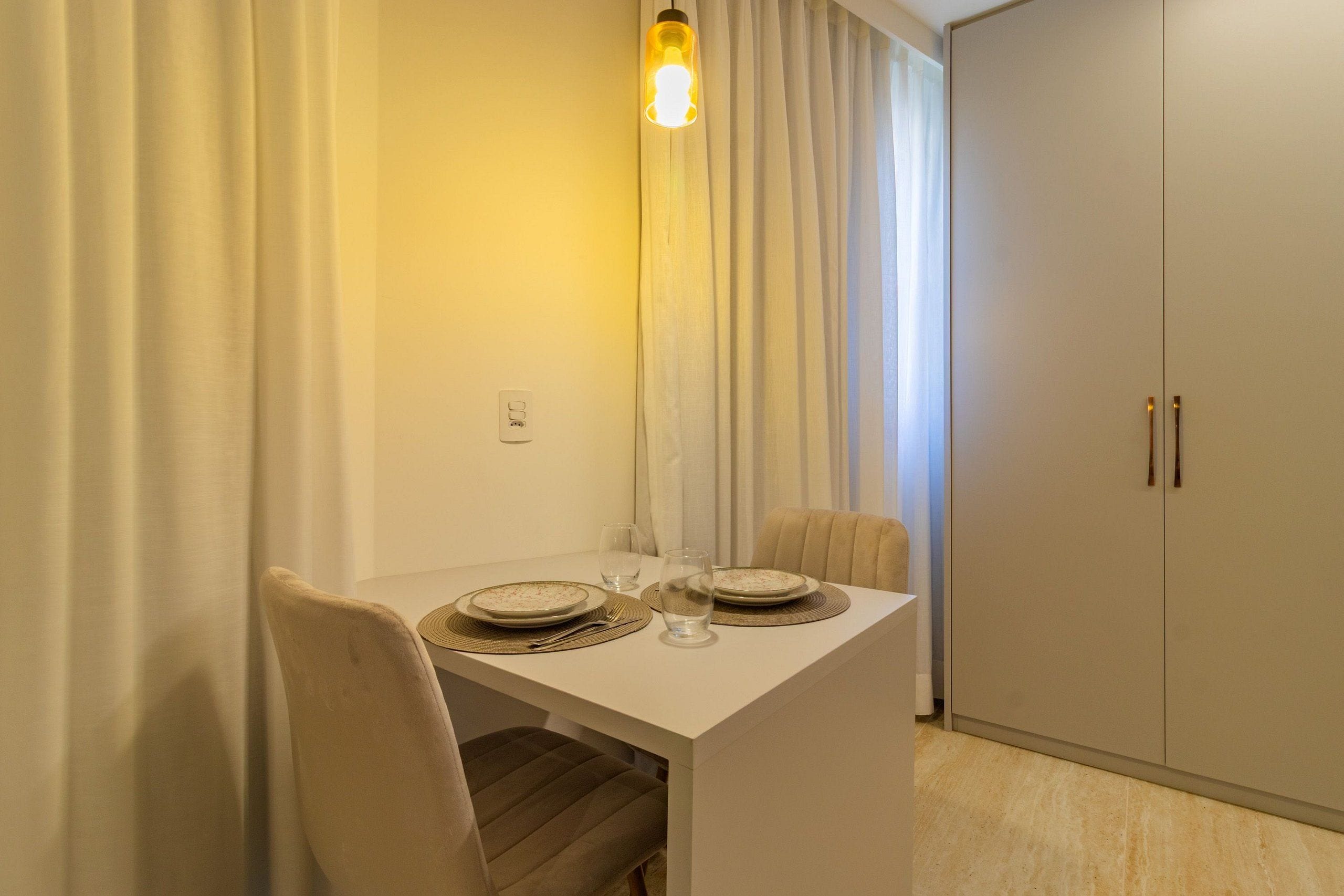 Un comedor con una mesa blanca dispuesta para dos, un colgador de luz y un armario de dos puertas.