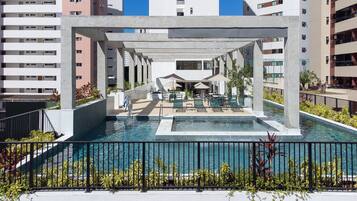 Área de piscina externa com espaço de estar coberto, cercada por uma grade de proteção e vasos de plantas.