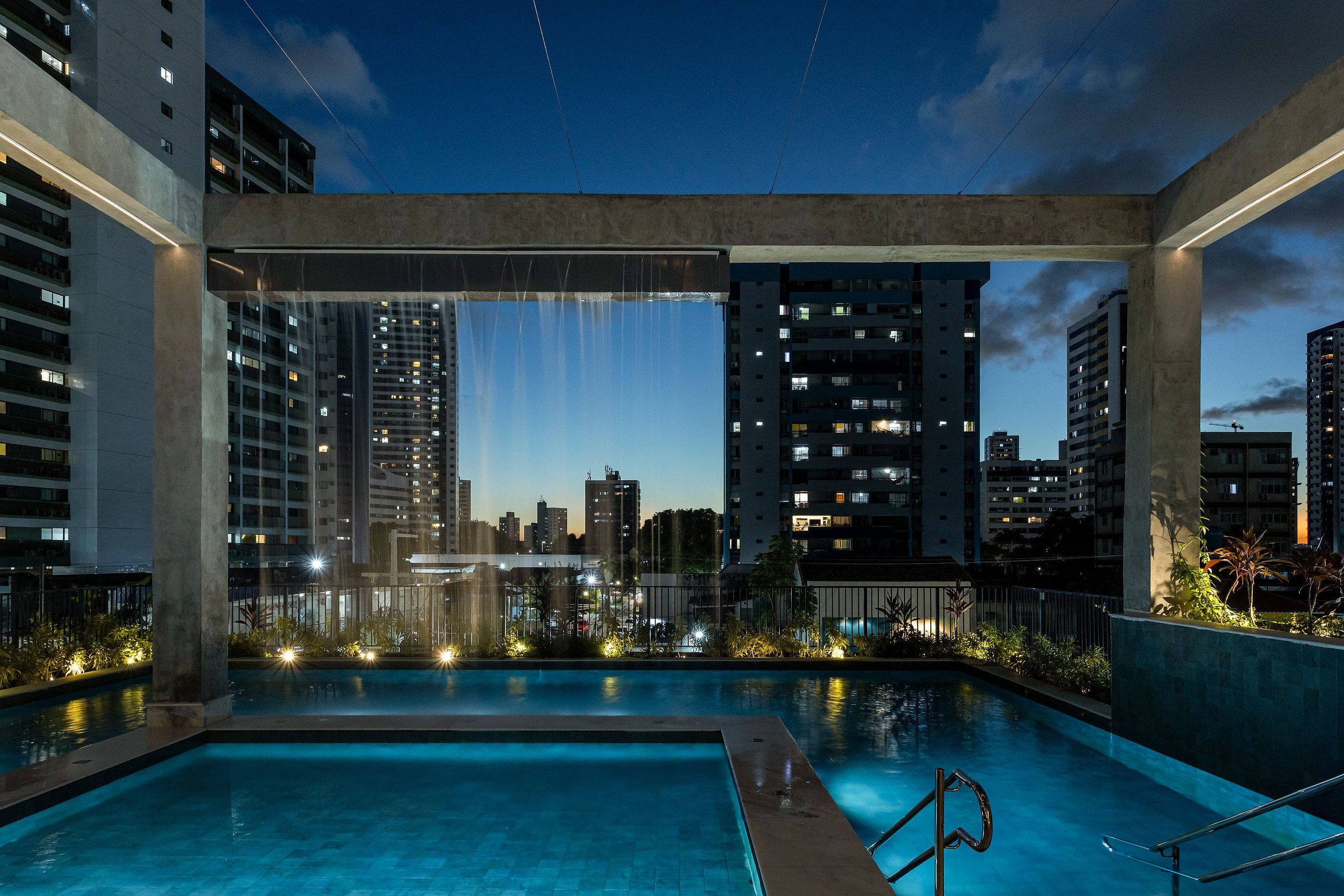 An outdoor swimming pool with a cityscape view, surrounded by high-rise buildings and illuminated by artificial lighting.