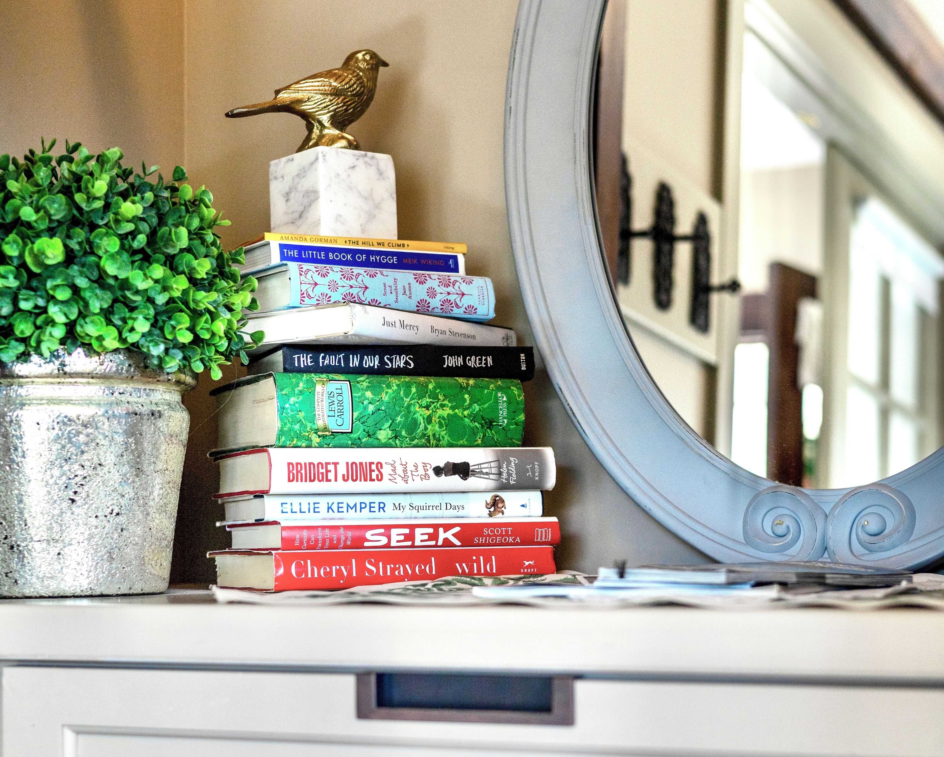 Een stapel boeken op een plank, met een decoratief vogelbeeldje en een potplant ernaast.