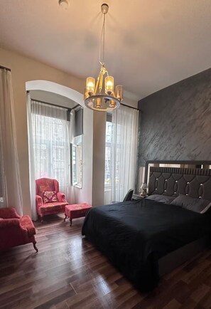 A hotel room with a large bed, a red armchair, a small table, and a chandelier.
