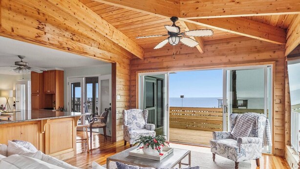 A spacious living area with a wooden ceiling, a ceiling fan, and a view of the ocean through large sliding glass doors.