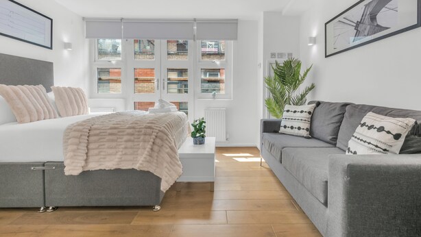 Une chambre à coucher moderne avec un lit, un canapé gris, une table de chevet blanche et une fenêtre donnant sur des immeubles.
