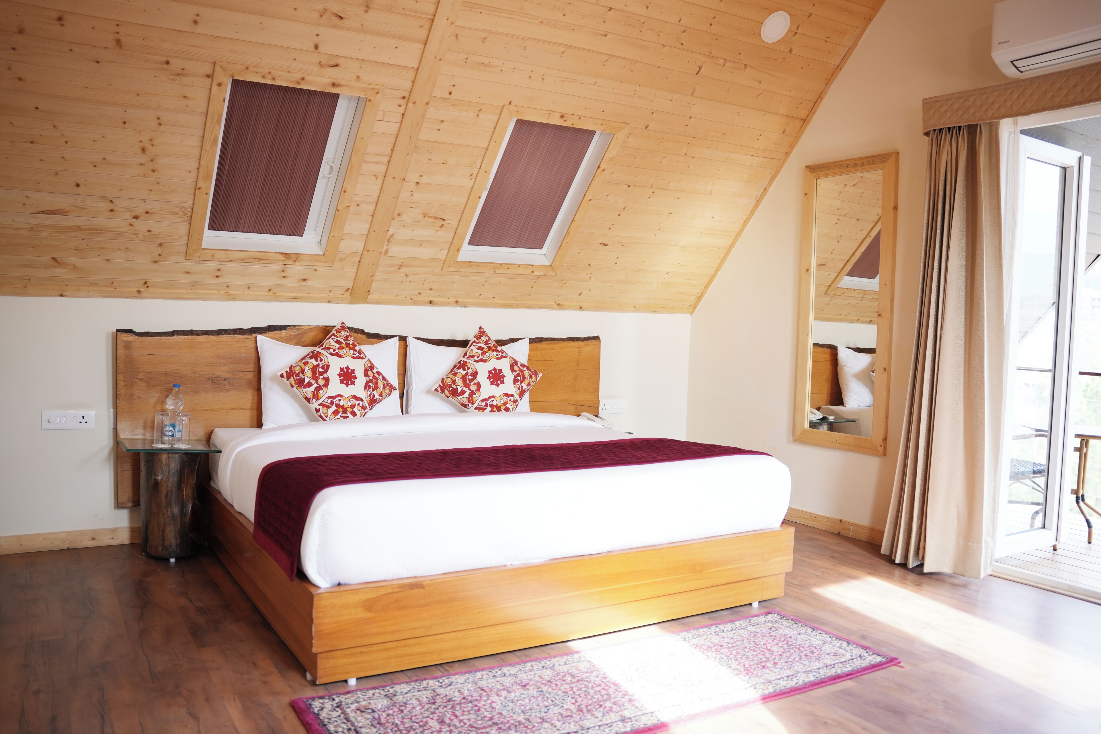 Une chambre avec un plafond en bois, des fenêtres de toit, un lit recouvert d’une couverture rouge et une table de chevet avec une bouteille d’eau.