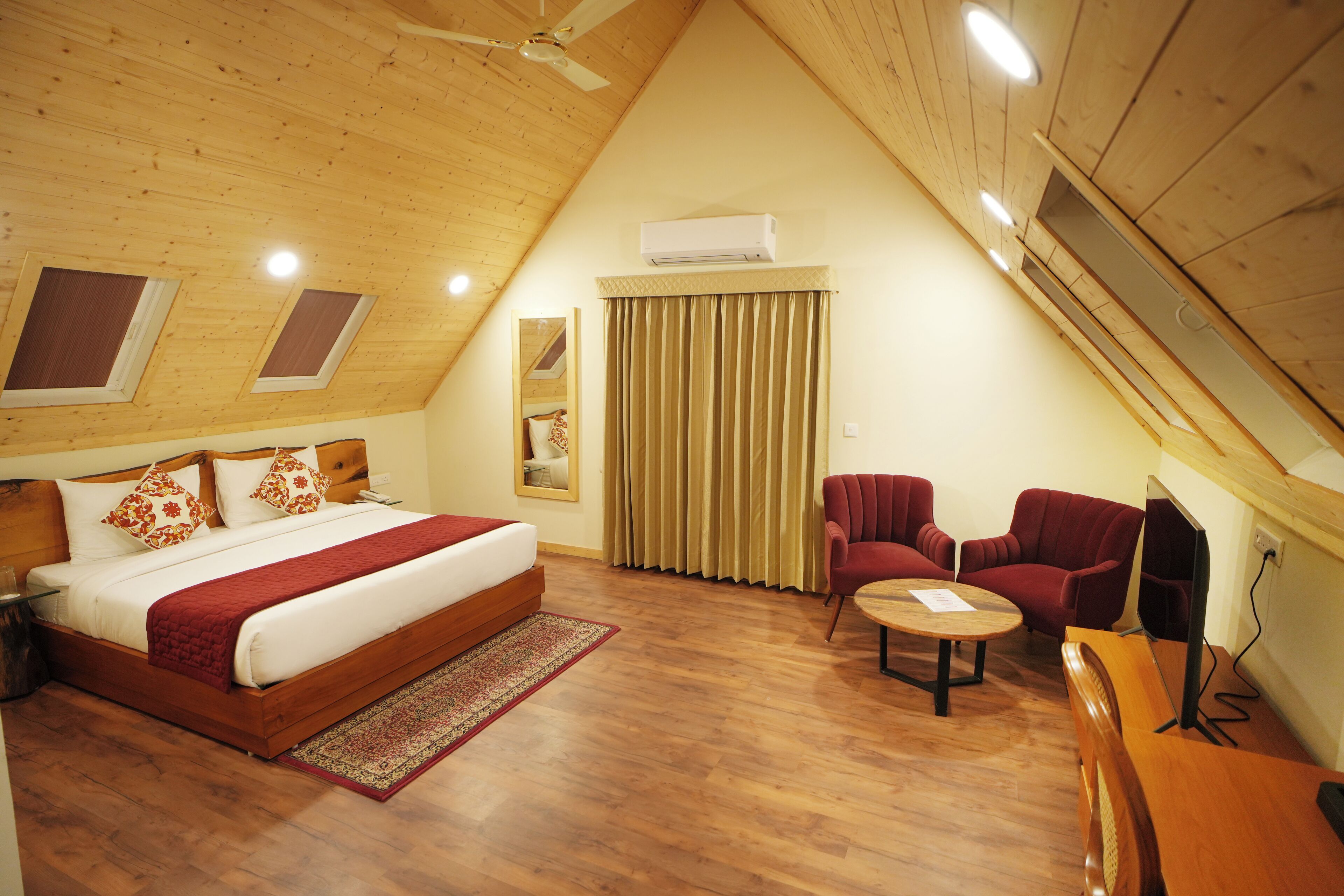 A bedroom with a wooden ceiling, a bed with a red blanket, two red chairs, a small round table, and a television.